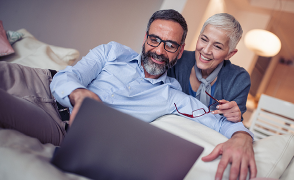 an image of an older couple sitting on a couch looking at their computer with the wife looking over the husbands shoulder holding glasses